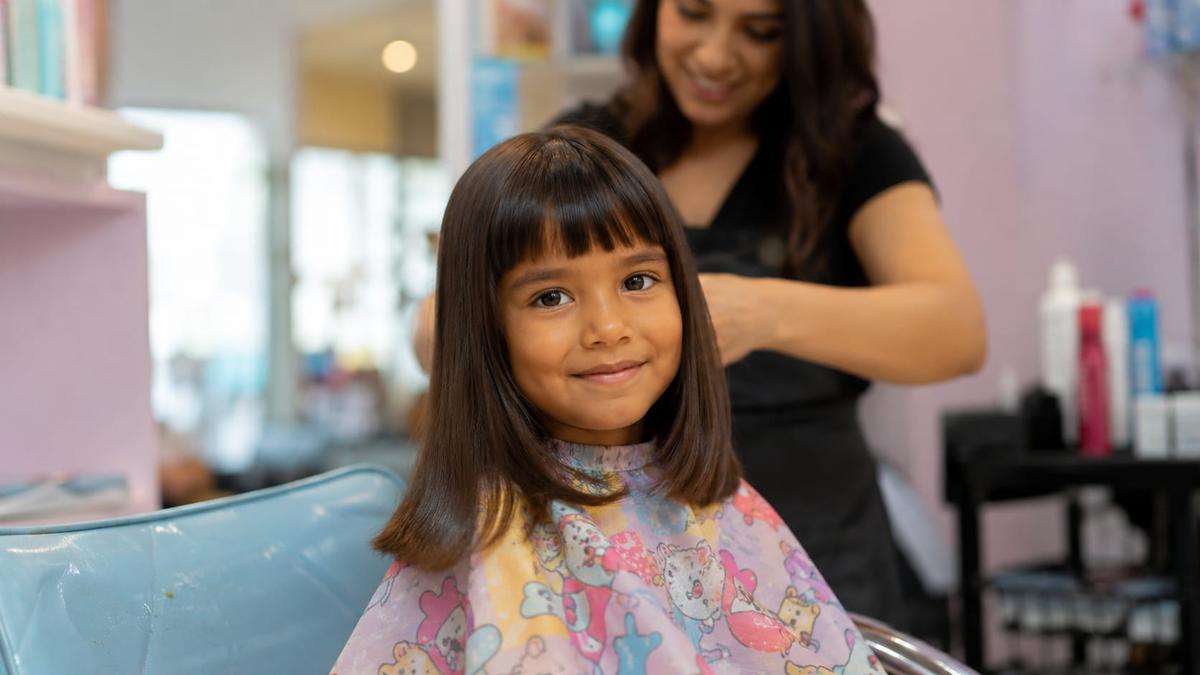 Criança sorridente com corte de cabelo chanel e franja reta clássica, vestindo uma capa colorida de personagens em um salão infantil.