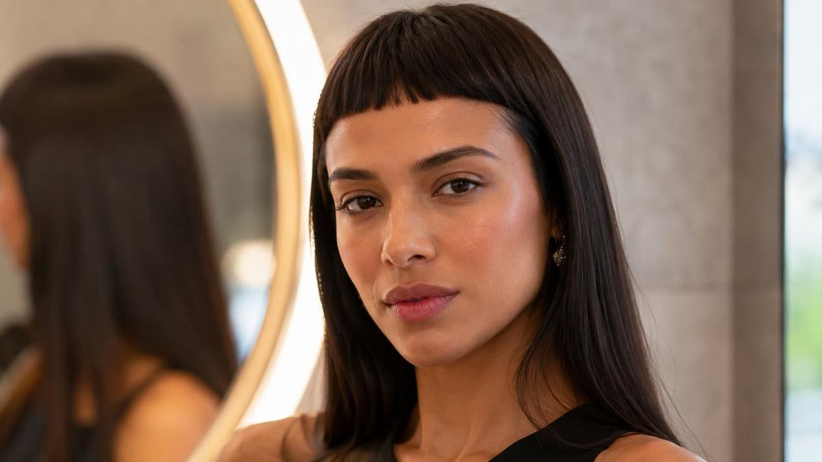 Close de mulher com franja baby bangs reta em cabelo longo liso, posando de perfil em frente ao espelho iluminado.