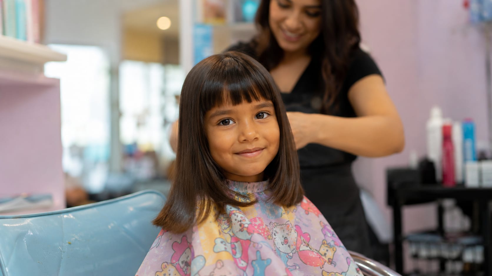 Criança sorridente com corte de cabelo chanel e franja reta clássica, vestindo uma capa colorida de personagens em um salão infantil.