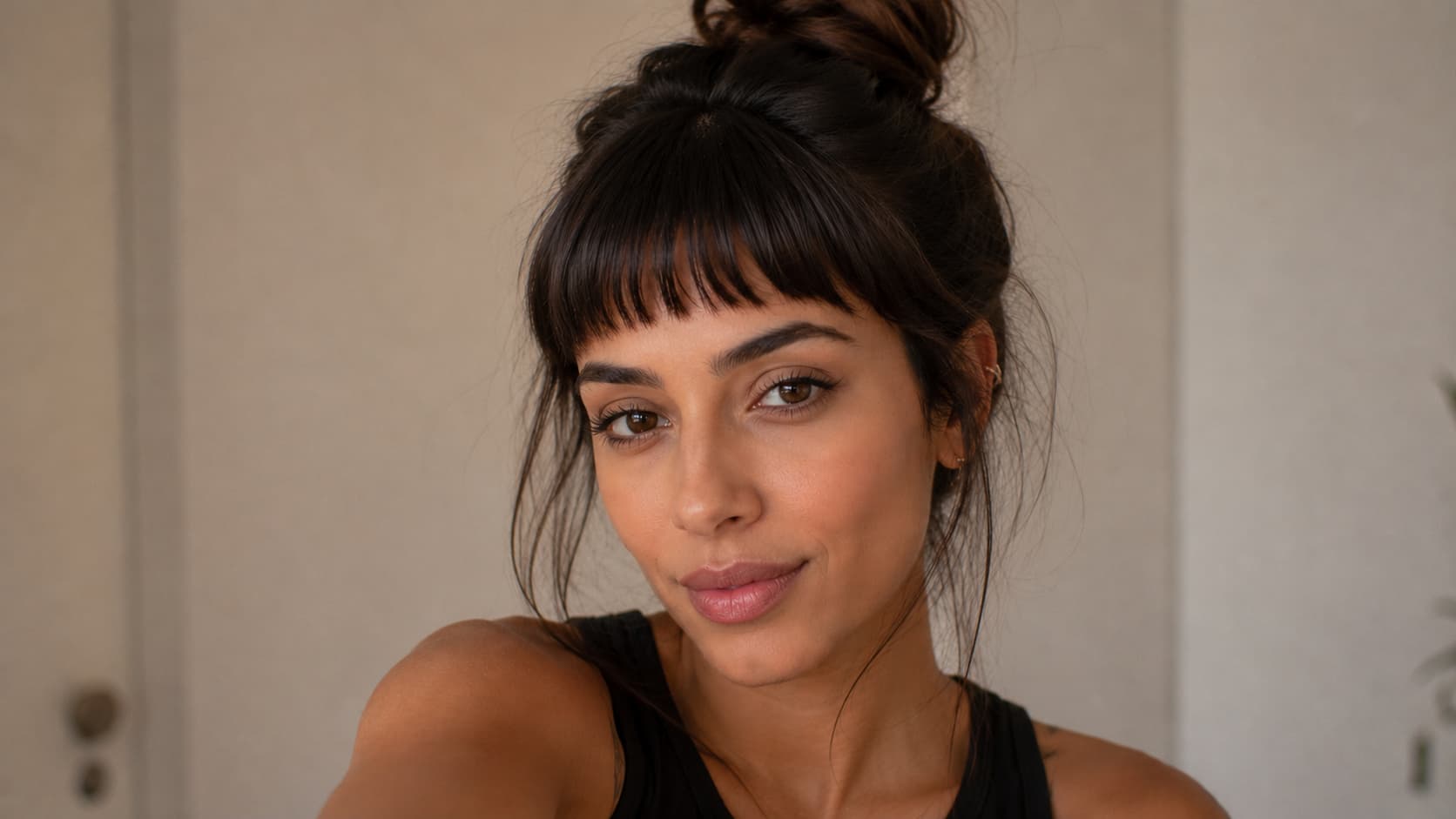 Penteado em coque alto com destaque para a franja curta no meio da testa desfiada em cabelo escuro. Mulher olha para a câmera.