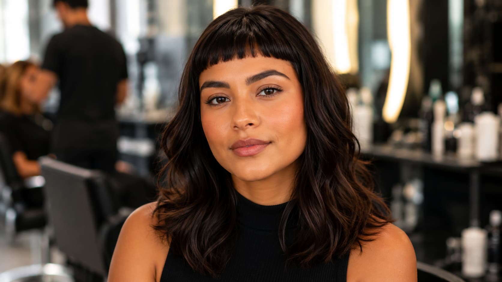 Mulher com rosto oval e cabelo médio ondulado escuro e franja curta reta sorri sutilmente em um salão de beleza.