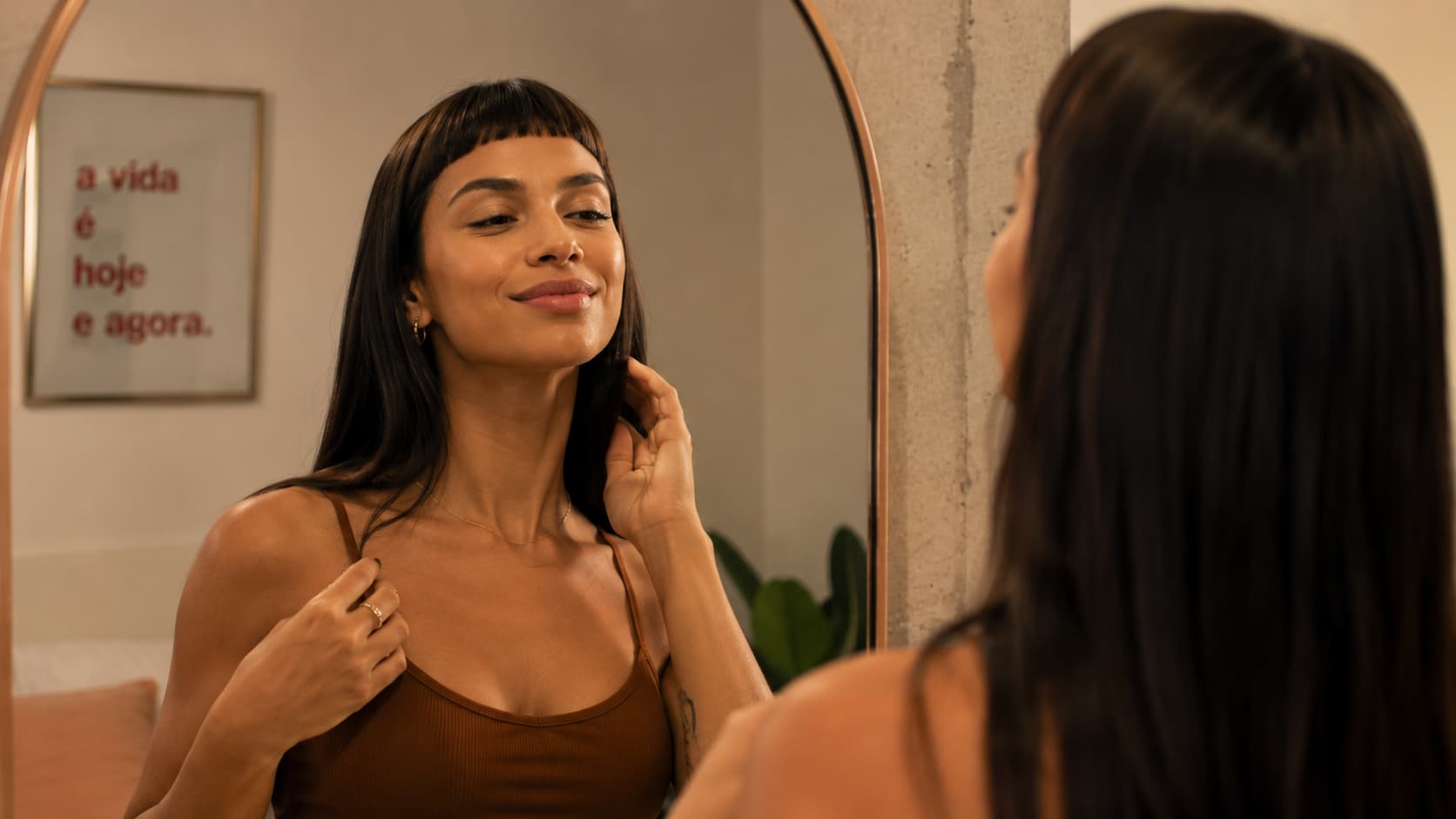 Mulher de cabelo longo e franja curta se olha no espelho. Quadro ao fundo diz a vida é hoje e agora.